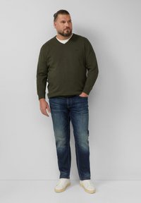 Maglione verde a V sopra una camicia bianca, abbinato a jeans in denim blu e sneakers bianche. L'uomo è in piedi contro uno sfondo neutro.