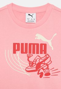 Camiseta de algodón rosa con un gran logo rojo de "PUMA" y un gráfico divertido de una zapatilla de deporte en estilo cartoon. Complementada con un gato de PUMA blanco.