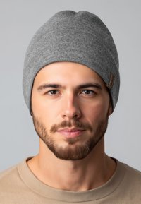 Grijze gebreide muts met een opgevouwen rand en leren merkembleem, gedragen door een persoon met korte baard en een shirt in neutrale kleuren, tegen een lichte achtergrond.