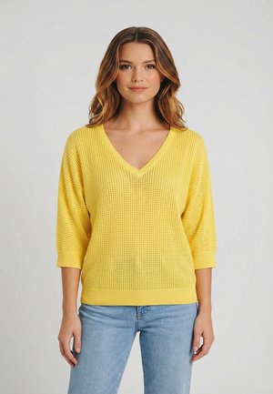 Donna che indossa un maglione giallo lavorato a maglia con scollo a V e maniche a tre quarti, e jeans azzurri, in piedi contro uno sfondo chiaro e uniforme.