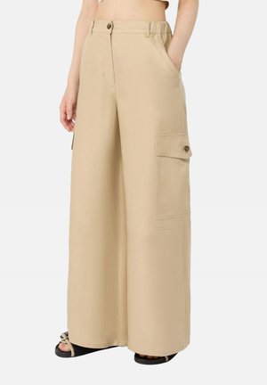 Pantaloni cargo beige a gamba larga con tasca con patta e bottone, bottone frontale e zip, indossati con sandali neri open toe.