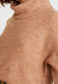 Gebreide coltrui in een zachte perzikkleur, met een losse pasvorm en een gestructureerde stof met subtiele toonvariaties.