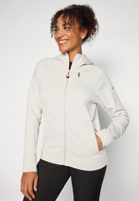 Haut à fermeture éclair beige clair avec un design ajusté, texture douce, capuche et détails de fermeture éclair roses. Associé à un pantalon noir.