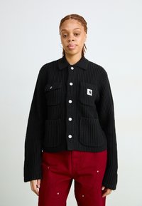 Schwarze gerippte Jacke mit Kragen, die zwei Fronttaschen und silberne Knöpfe hat. Trägt man über roten Hosen, die aus einem strukturierten Strickmaterial bestehen.