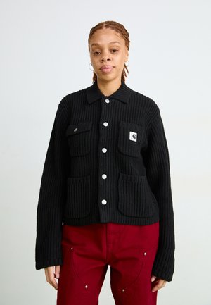 Schwarze gerippte Jacke mit Kragen, die zwei Fronttaschen und silberne Knöpfe hat. Trägt man über roten Hosen, die aus einem strukturierten Strickmaterial bestehen.