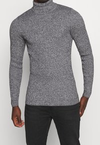 Man som bär en åtsittande grå och svart melerad polotröja tillsammans med svarta byxor, står framför en enkel vit bakgrund.