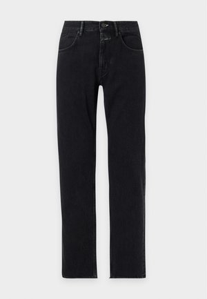Homme portant une veste marron boutonnée sur une chemise blanche, un pantalon noir et des mocassins noir et blanc, debout devant un fond uni.