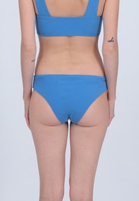 Bas de bikini bleu avec une texture lisse, coupe basse et couverture minimale, vu de dos. Pas de motifs ou d'accents visibles.