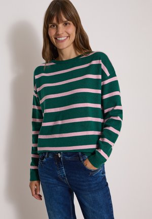 Femme souriante portant un pull vert avec des rayures horizontales roses et un jean bleu, une main dans la poche, debout devant un fond uni.