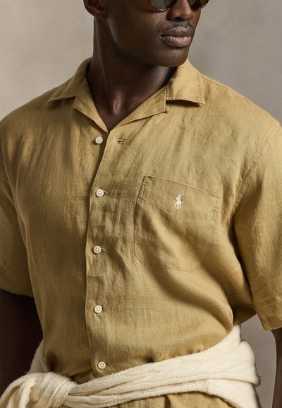 Mann trägt ein senfgelbes kurzärmeliges Hemd mit Knöpfen, mit einem kleinen weißen, auf der Brusttasche gestickten Logo, und einen cremefarbenen Pullover um die Taille gebunden.