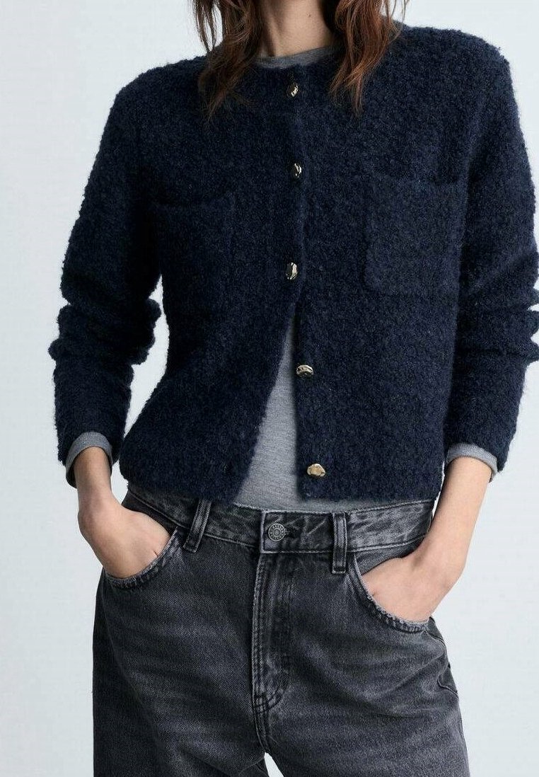 Femme portant un cardigan duveteux bleu marine boutonné par-dessus une chemise grise, les mains dans les poches d'un jean noir délavé.