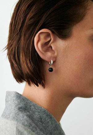 Rövid barna haj a fül mögött, ezüst karika fülbevalóval, amelyen egy kis fekete gyöngy található, szürke ruhakabát gallérját viseli.