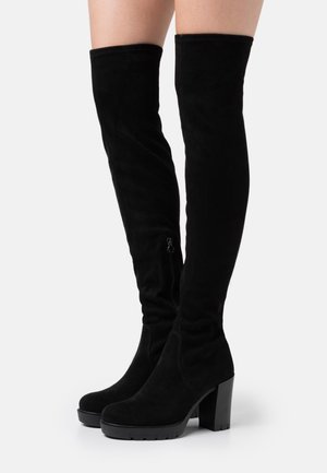 Bottes en daim noir au-dessus du genou avec talons épais et fermetures éclair latérales, portées sur des jambes à peau claire, posées sur une surface blanche.