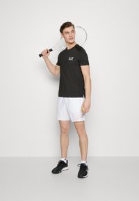 Camiseta deportiva negra con logo de EA7, pantalones cortos blancos con acentos naranjas, zapatillas deportivas negras, sosteniendo una raqueta de tenis. Fondo neutro.