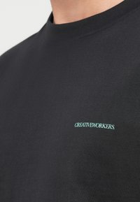 Κοντινό πλάνο μαύρου T-shirt με στρογγυλή λαιμόκοψη και μικρό ανοιχτό μπλε κείμενο "CREATIVEWORKERS." στην αριστερή πλευρά του στήθους.