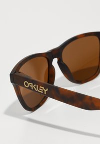 Ochelari de soare din scoică cu lentile maro, având un logo auriu proeminent "OAKLEY" pe braț, cu o finisare netedă și lucioasă.