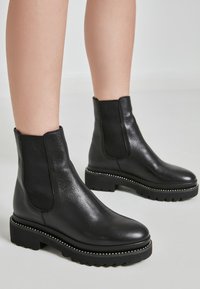 Stivaletti neri in pelle con pannelli elastici laterali, finitura testurizzata e suole in gomma spessa con borchie decorative argento lungo il bordo.