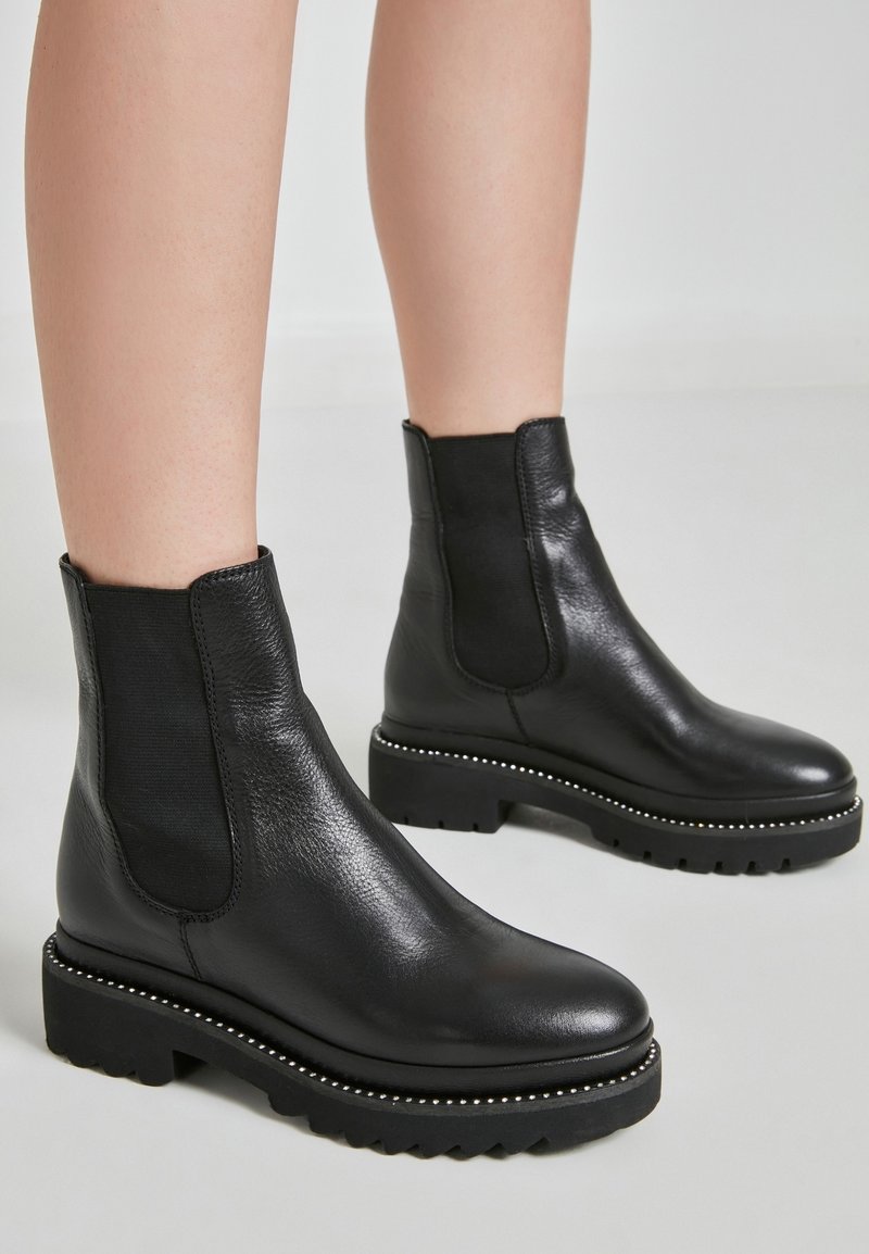 Stivaletti neri in pelle con pannelli elastici laterali, finitura testurizzata e suole in gomma spessa con borchie decorative argento lungo il bordo.