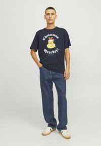 Marineblå bomulds T-shirt med et gult gummiand grafik med rensdyrhorn og tekst, parret med løse mørkeblå jeans og hvide sneakers.