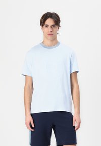 Tricou din bumbac de culoare albastră deschisă, cu un logo circulat texturat pe față și un guler cu dungi ribbed. Asortat cu pantaloni scurți bleumarin.