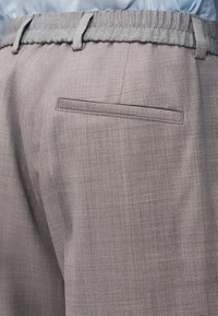 Gros plan sur une personne portant un pantalon habillé gris avec une poche arrière cintrée et une taille élastique, assorti d'une chemise bleu clair.