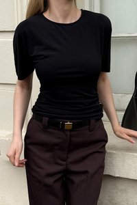 Juodas trumpomis rankovėmis viršus, turintis lygią tekstūrą ir prigludusią išvaizdą, derinamas su aukštu liemeniu rudomis kelnėmis ir juodu diržu su auksiniu užsegimu.