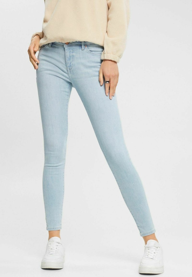 Personne portant un jean skinny bleu clair, un pull beige en laine pelucheuse, des baskets blanches et une bague noire au doigt annulaire gauche, debout les jambes écartées.