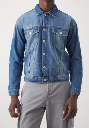 Homme portant une veste en jean bleue boutonnée avec des poches à rabat et un pantalon gris, debout devant un fond clair uni.