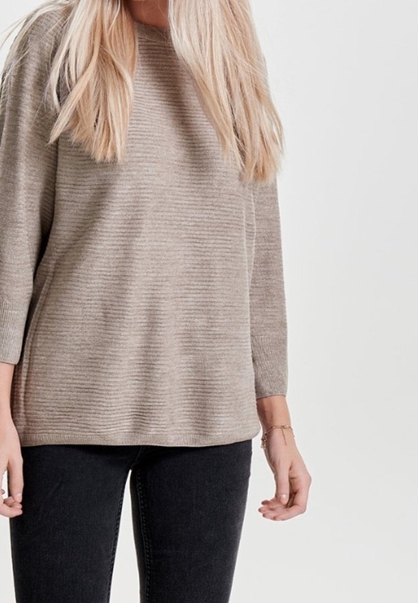 Femme aux cheveux longs et blonds portant un pull côtelé beige et un jean noir sur un fond clair uni.