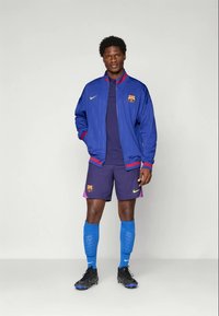 Blå og rød trackjakke med striber, lilla shorts, blå sokker og sorte fodboldstøvler, med et holdlogo på brystet og shortsene.