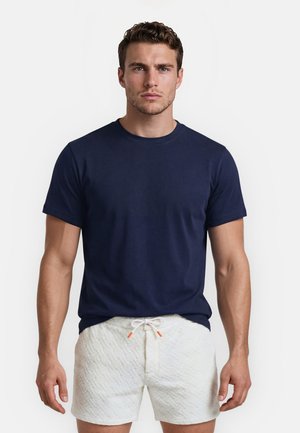 Mand stående iført en marineblå T-shirt med korte ærmer og hvide teksturerede shorts med snor mod en ensfarvet hvid baggrund.