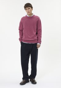 Rózsaszín kötött pulóver kerek nyakkivágással, sötétkék kordbársony nadrággal és barna cipővel párosítva. A háttér egyszerű fehér.