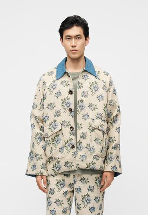 Jaunas vyras dėvi smėlio spalvos švarką su gėlių raštais ir derančias kelnes, stovintis priešais paprastą baltą foną.