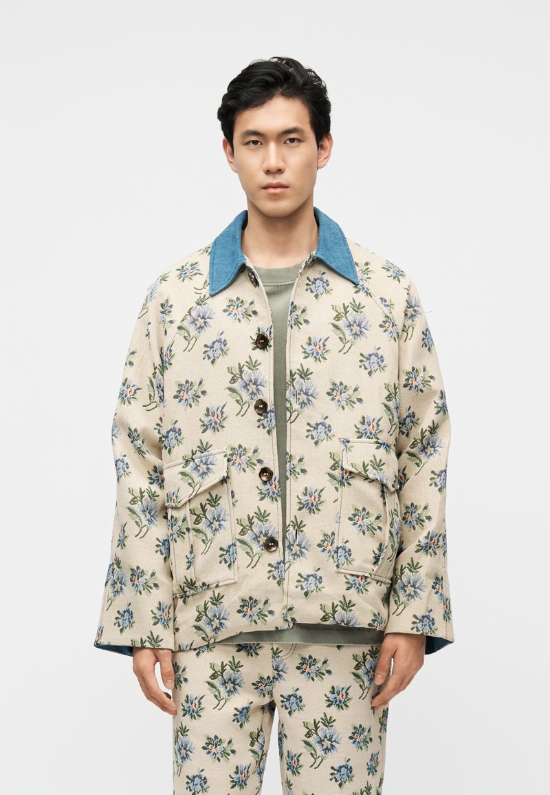 Νεαρός άνδρας με μπεζ σακάκι με λουλουδάτο σχέδιο και ταιριαστό παντελόνι, στέκεται μπροστά σε απλό λευκό φόντο.