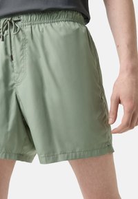 Zelené plavkové šortky s elastickým pasem, stahovací šňůrkou a bočními kapsami. Vyrobené z lehkého, hladkého materiálu s matným povrchem.