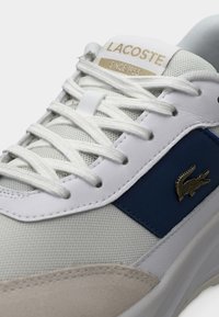 Αθλητικά παπούτσια Lacoste με λευκό επάνω μέρος από δίχτυ, μπλε δερμάτινες λεπτομέρειες, μπεζ σουέντ διακοσμήσεις και χρυσό λογότυπο στη γλώσσα. Κορδόνια με υφή.