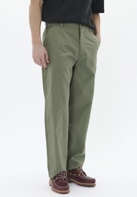 Pantaloni subțiri de culoare verde măslin, cu croială dreaptă, dotate cu buzunare laterale și fermoar, asortați cu pantofi din piele maro.
