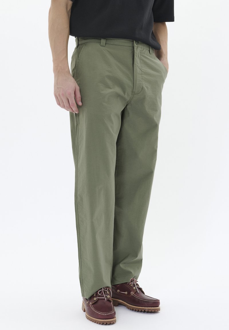 Pantaloni subțiri de culoare verde măslin, cu croială dreaptă, dotate cu buzunare laterale și fermoar, asortați cu pantofi din piele maro.
