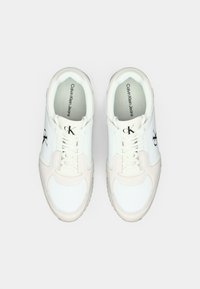 Witte sneakers met lichtbeige accenten, vervaardigd van gladde leren en textiele materialen, met veters en een zwart "ck"-logo aan de zijkant.