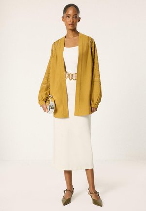 Femme debout dans une robe midi blanche avec une ceinture dorée, une veste ouverte jaune moutarde avec des manches ornées, et des talons pointus olive.