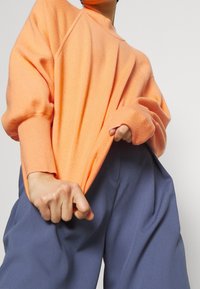 Suéter de punto naranja de corte holgado, con puños y escote acanalados, combinado con pantalones anchos azules. Textura suave y silueta relajada.