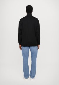 Giacca nera con colletto alto e orlo elasticizzato, vista dal retro. Abbinata a jeans in denim azzurri e sneakers bianche.