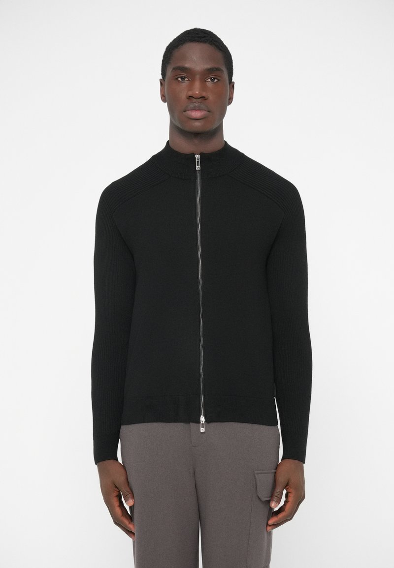 Čierny zapínací sveter so zipsom, rebrovanou textúrou, vysokým golierom a dlhými rukávmi; hladká látka s kovovými detailmi na zips a priliehavým strihom.