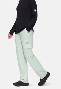 Ljus mintgrön zip-off-byxor med fickor och en cirkulär logotyp, i kombination med en svart långärmad tröja och svarta vandringsskor.