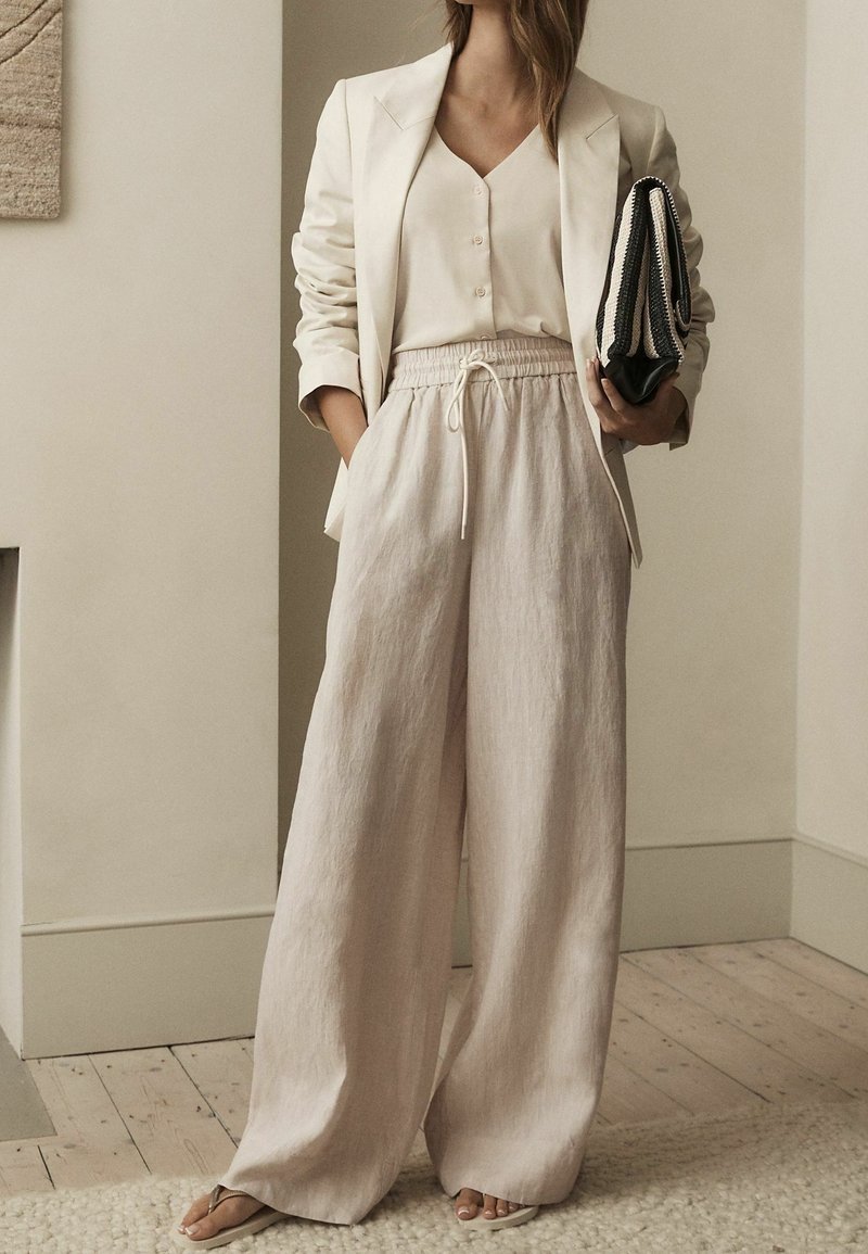 Femme portant un blazer beige, un chemisier crème, un pantalon large en lin, tenant une pochette noir et blanc dans une pièce aux tons neutres avec un sol en bois.