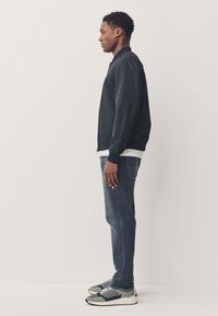 Homme debout de profil, portant une veste bomber noire, une chemise blanche, un jean foncé et des baskets grises sur un fond clair uni.