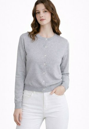 Femme aux cheveux bruns ondulés portant un cardigan gris clair boutonné et un pantalon blanc, debout avec une main dans la poche sur fond blanc.