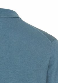 Zbliżenie niebieskiego dzianego materiału, pokazujące szew ramienia i brzeg kołnierza odzieży na białym tle.