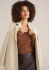 Beige Wollmantel mit Reverskragen, kombiniert mit einem braunen Langarmtop und einem braunen Lederrock, der glatte Texturen präsentiert.