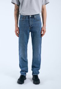 Vaqueros de mezclilla azul con un diseño de pierna recta, que cuentan con cinco bolsillos, un cierre de botón y una textura desgastada. Llevados con zapatillas oscuras.